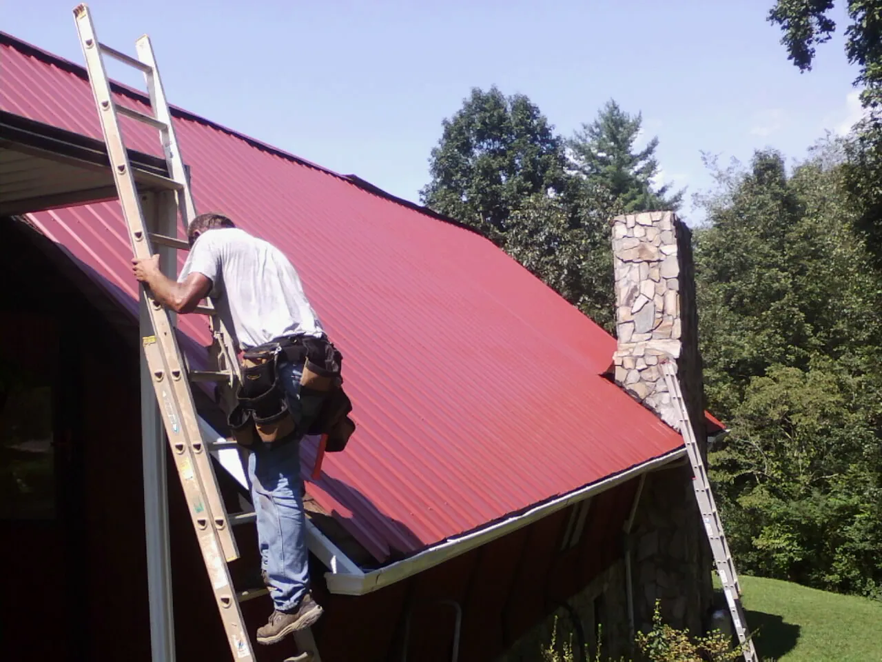 Roofer on a ladder beside a red metal roof and stone chimney — Soffit & Fascia Repair by Roofing Brothers in Lucama